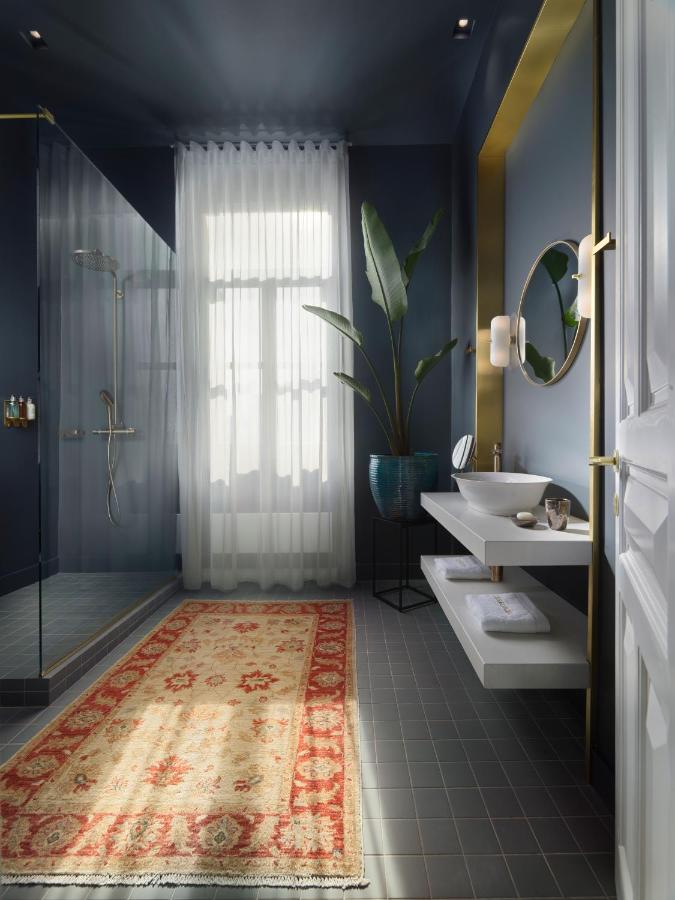 A modern bathroom reminiscent of the best boutique hotels in Athens, with dark walls, a walk-in glass shower, a large window with sheer curtains, a floating sink and round mirror, potted plant, and a patterned red and gold rug on gray tile floor.
