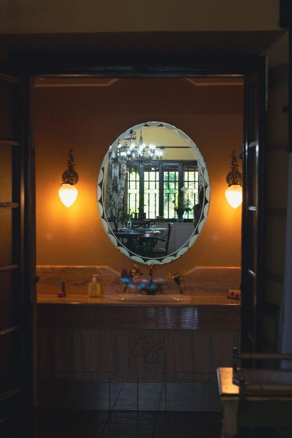 A warmly lit bathroom with an oval mirror above a marble counter, often found in elegant hotels in Cantabria. Two wall lamps flank the mirror, reflecting a chandelier and large windows with lush greenery outside.