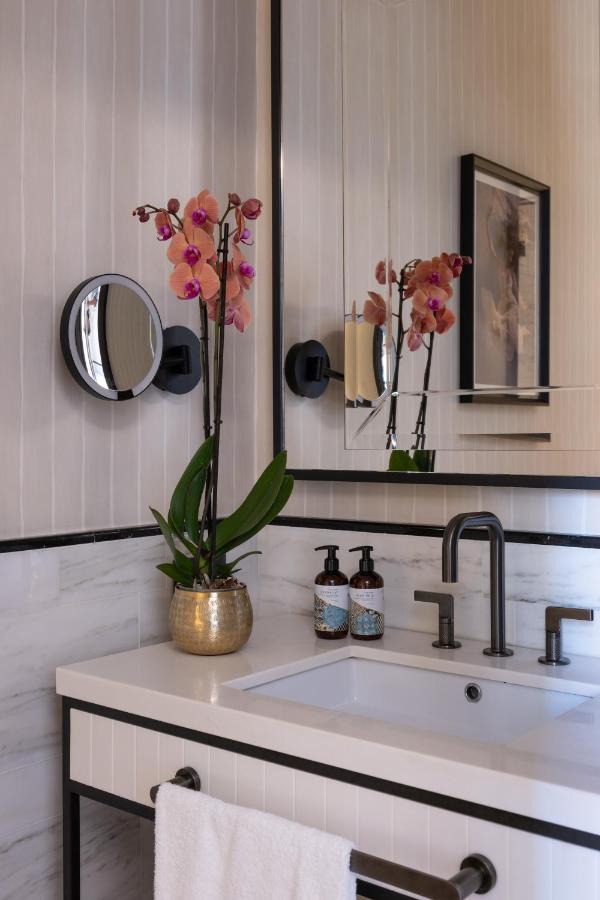 13 UNIQUE Boutique Hotels in Rome 7 Modern bathroom sink like those in boutique hotels in Rome, Italy, with a brushed metal faucet, white countertop, gold pot with pink orchid, two soap dispensers, wall-mounted mirror, hanging towel, and framed art reflected above.
