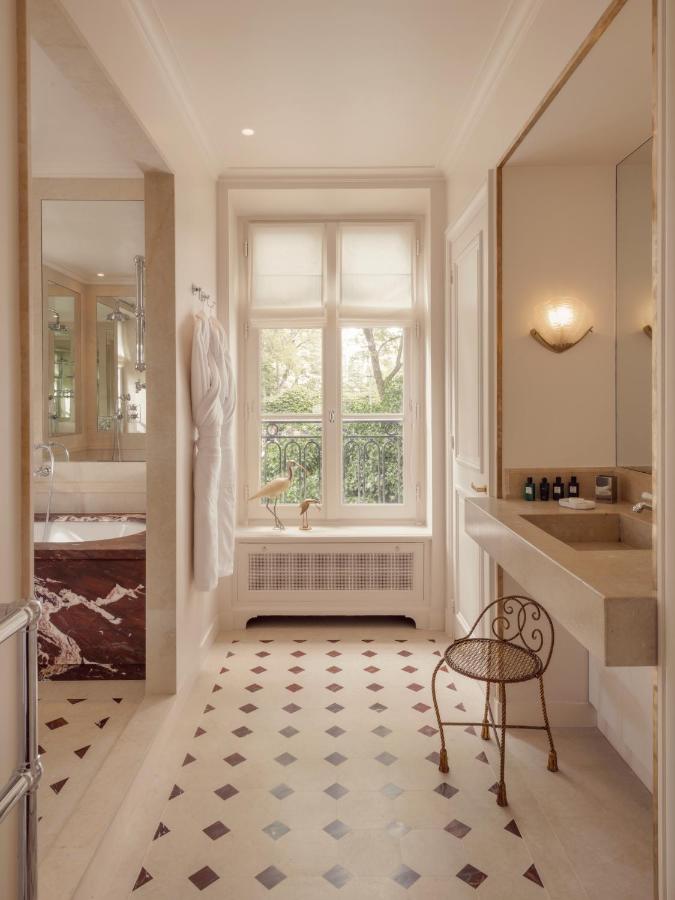 13 Unique Hotels in Paris for a Stylish Stay 12 Elegant bathroom at one of the unique hotels in Paris, featuring a large window, beige countertop and sink, small metal chair, wall-mounted mirror, fresh towels, and a bathtub with brown marble accents atop diamond-patterned tile flooring.