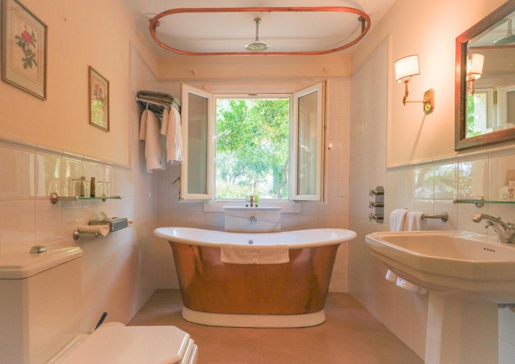 TOP 10 Luxury Hotels in Andalucía 41 A bright bathroom reminiscent of luxury hotels in Andalucía, featuring a copper bathtub beneath an open window, white sink, toilet, wall-mounted towel rack with robes, and framed art on the tiled walls. Green trees are visible outside.