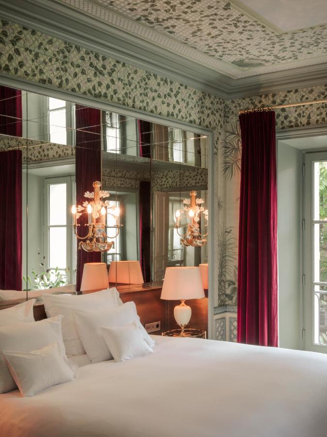 13 Unique Hotels in Paris for a Stylish Stay 11 Elegant bedroom with patterned wallpaper, large mirrors, and a chandelier. Reminiscent of unique hotels in Paris, the bed with white linens sits by a wooden nightstand and lamps. Tall windows are adorned with deep red velvet curtains.