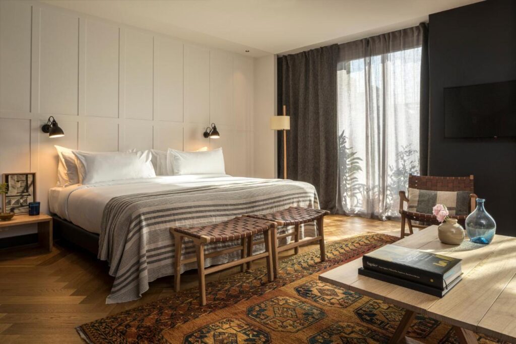 13 Unique Hotels in Barcelona, Spain 6 A modern hotel room in one of the unique hotels in Barcelona, featuring a large bed with striped bedding, bedside tables with lamps, woven benches, an ornate rug, stylish decor on a table, wooden flooring, and large windows with sheer and dark curtains.