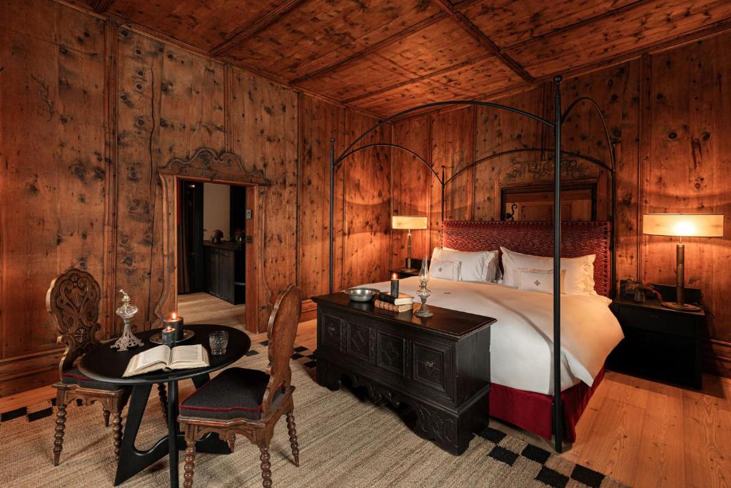 A cozy, rustic bedroom inspired by the best hotels in the Dolomites, with wood-paneled walls and ceiling, a large canopy bed with white linens, a dark wooden chest at the foot, and a round table with books. Warm lighting fills the room.