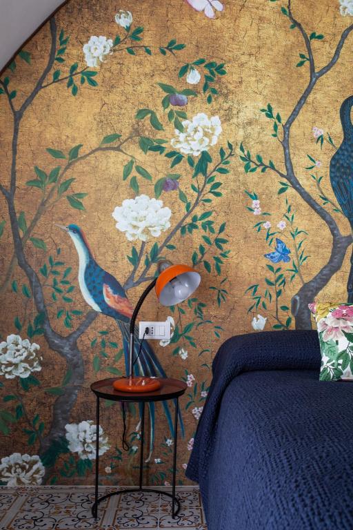 7 Best Hotels in the Aeolian Islands 12 A bedroom in the style of hotels in the Aeolian Islands, with a gold and floral mural featuring birds on the wall, a navy blue bedspread, a table with an orange lamp, and patterned floor tiles.