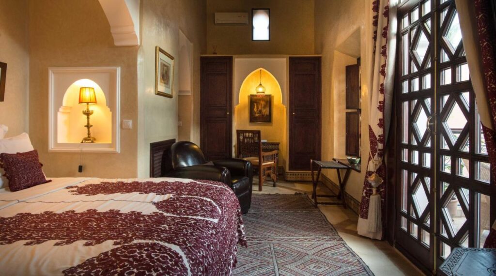 10 Unique Riads in Marrakech with Pool 5 A cozy bedroom in the style of riads in Marrakech with pool, featuring a large bed with red-patterned bedding, a leather armchair, small table and chairs, a lamp in a wall niche, and tall windows with patterned curtains letting in natural light.