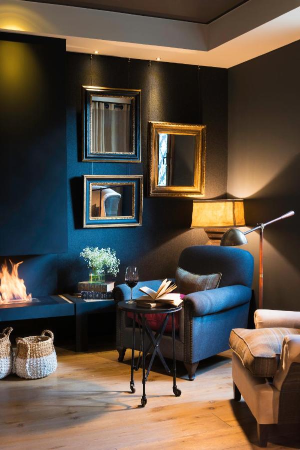 13 Unique Hotels in Barcelona, Spain 12 A cozy living room reminiscent of unique hotels in Barcelona, with a dark accent wall, gold-framed mirrors, a lit fireplace, a gray armchair with an open book and wine glass on a small table, and a basket on the wooden floor.