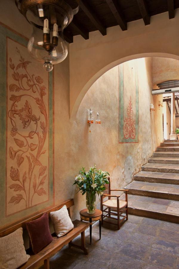 A rustic hallway reminiscent of boutique hotels in Granada, with stone steps, floral wall murals, wooden benches with cushions, a chair, a small table holding a vase of flowers, and a glass light fixture hanging from a wooden ceiling.