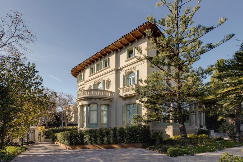 13 Unique Hotels in Barcelona, Spain 34 A large, elegant beige villa with tall windows, balconies, and a red-tiled roof stands among tall trees and green shrubs—reminiscent of unique hotels in Barcelona—its driveway leading to an entrance shaded by lush foliage on a sunny day.