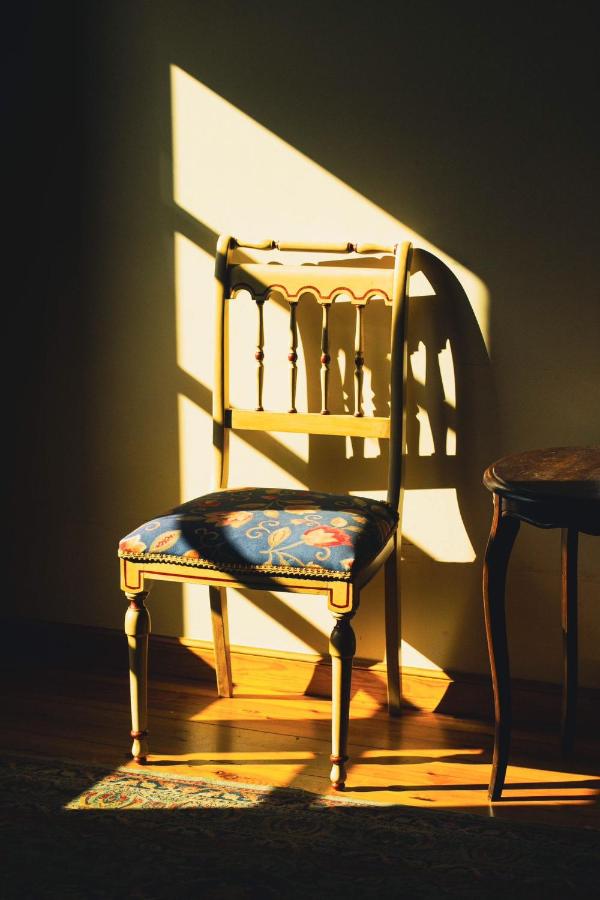 A vintage wooden chair with a patterned cushion sits in sunlight streaming through a window, casting strong shadows on the wall and floor—evoking the charm of boutique hotels in Cantabria—beside a small round wooden table.