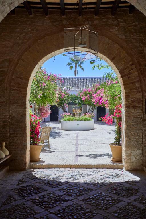TOP 10 Luxury Hotels in Andalucía 17 View through a stone archway into a sunlit courtyard with flowering plants, potted trees, a central white fountain, and wicker chairs—evoking the serene elegance of luxury hotels in Andalucía—under a blue sky and palm trees.