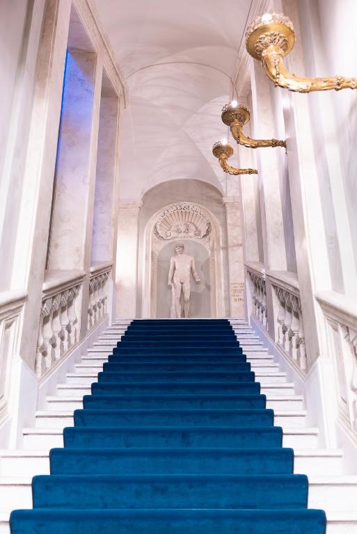 13 UNIQUE Boutique Hotels in Rome 53 A grand white marble staircase with a royal blue carpet runner leads to a classical nude statue in an arched alcove—evoking the refined elegance often found in boutique hotels in Rome, complete with ornate gold wall sconces and tasteful decor.