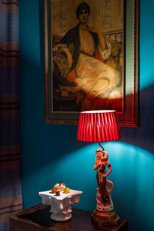 13 UNIQUE Boutique Hotels in Rome 54 A decorative lamp with a red shade stands on a small table beside a white pedestal of desserts, evoking the charming ambiance found in boutique hotels in Rome. A framed portrait of a seated woman hangs on the teal wall behind them.