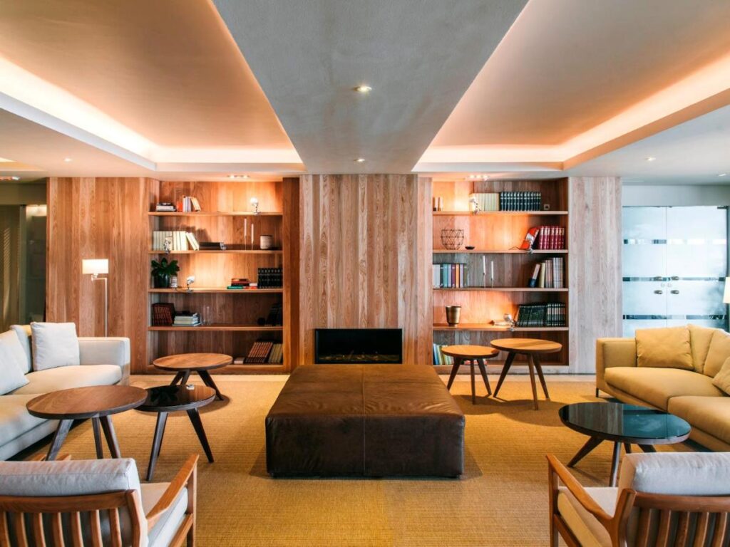 A modern lounge with cozy sofas, wooden bookshelves filled with books, round coffee tables, and warm lighting—reminiscent of elegant hotels in Cantabria. A large brown ottoman sits at the center of the room on a tan carpet.