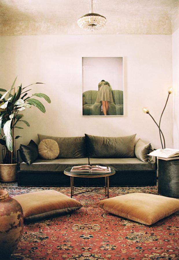 A cozy living room inspired by the best boutique hotels in Athens, featuring a green velvet sofa, decorative pillows, a round coffee table with an open book, large floor cushions on a patterned rug, a tall plant, and wall art above the sofa.