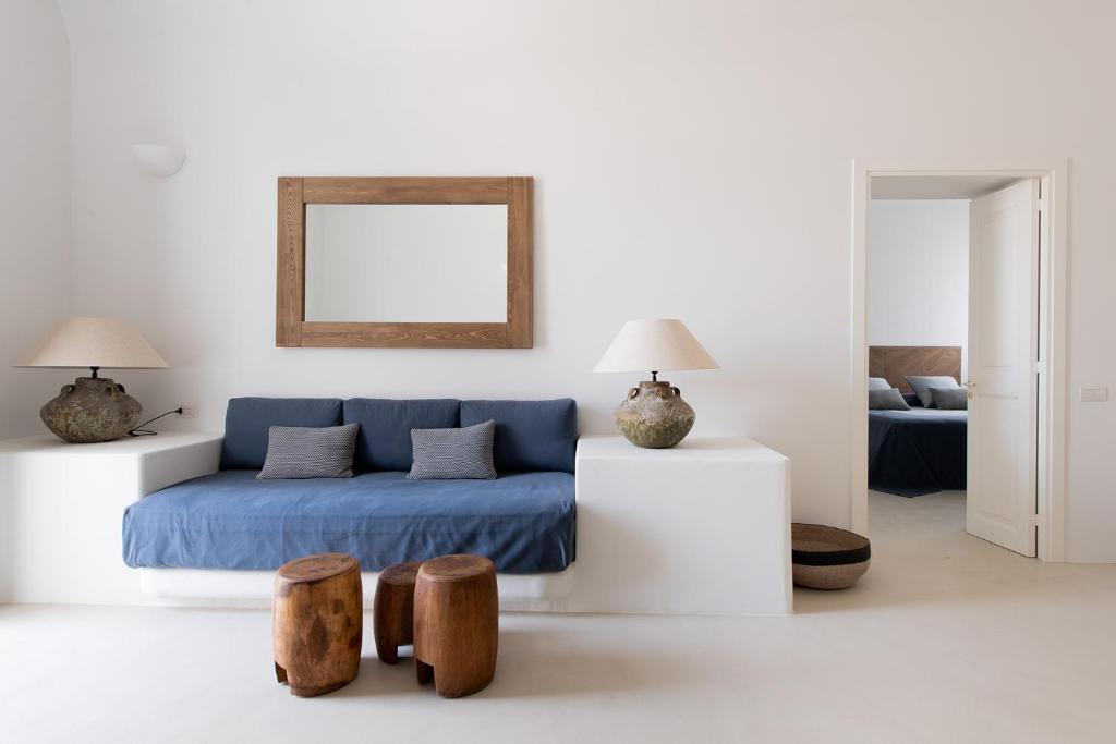 7 Best Hotels in the Aeolian Islands 4 A minimalist living room with a blue sofa, gray cushions, two wooden stools, large table lamps, and a wooden-framed mirror evokes the serene elegance found in the best hotels in the Aeolian Islands. An open door leads to a tranquil bedroom with a bed.