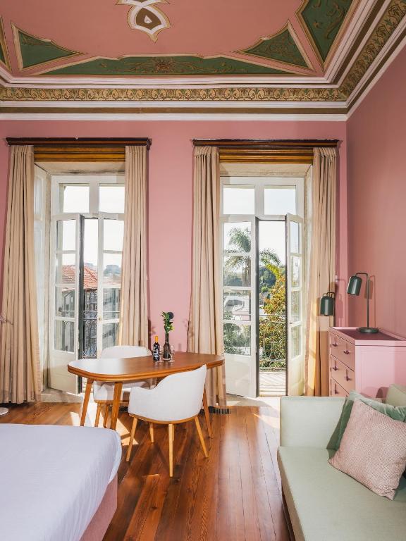 Top 12 Boutique Hotels in Porto, Portugal 29 A stylish room inspired by boutique hotels in Porto, with pink walls and ceiling, tall windows with beige curtains, a round wooden table with white chairs, a pink dresser, and a light green sofa. Sunlight streams in, revealing palm trees outside.