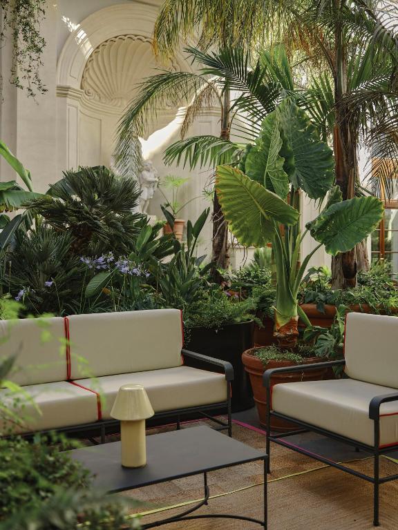 13 UNIQUE Boutique Hotels in Rome 49 A bright indoor garden with lush green plants, palm trees, and potted foliage surrounds modern beige cushioned chairs—reminiscent of the elegant lounges found in boutique hotels in Rome—centered around a black coffee table and yellow lamp under an arched feature.