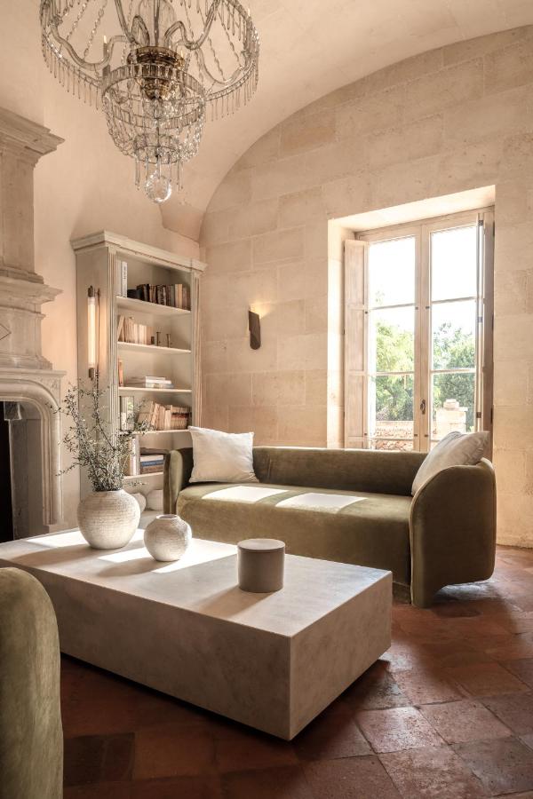 Unique Boutique Hotels in Menorca 5 Sunlit living room with arched ceiling, olive green sofas, a white rectangular coffee table with vases, built-in bookshelf, chandelier, and a window overlooking greenery—evoking the relaxed elegance of boutique hotels in Menorca. Warm, neutral tones throughout.