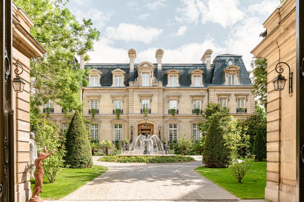 13 Unique Hotels in Paris for a Stylish Stay 33 Elegant three-story mansion with tall windows and a mansard roof, viewed from an open gate. Reminiscent of unique hotels in Paris, it features a circular driveway and fountain in front, surrounded by manicured gardens and statues.