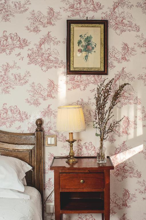 13 Unique Hotels in Paris for a Stylish Stay 44 A vintage bedside table with a brass lamp and a vase of dried flowers stands next to a wooden bed, evoking the timeless charm often found in unique hotels in Paris, against wallpaper with red patterns and a framed botanical print above.