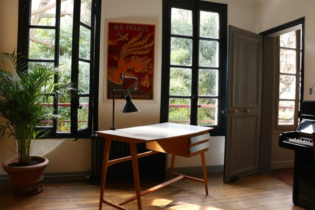 13 Unique Hotels in Paris for a Stylish Stay 16 A minimal home office with a wooden desk, black lamp, potted plant, and sunlight streaming through open windows. A vintage Air France poster, reminiscent of unique hotels in Paris, hangs on the wall beside an open door and a black piano.
