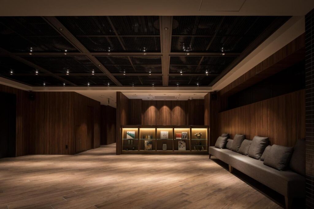 7 Best Hotels in Hiroshima, Japan 14 A modern, dimly lit lounge with wooden walls and floors, a long gray cushioned bench with pillows on the right, and a display case with books and decor illuminated by spotlights—an ambiance found in some of the best hotels in Hiroshima, Japan.