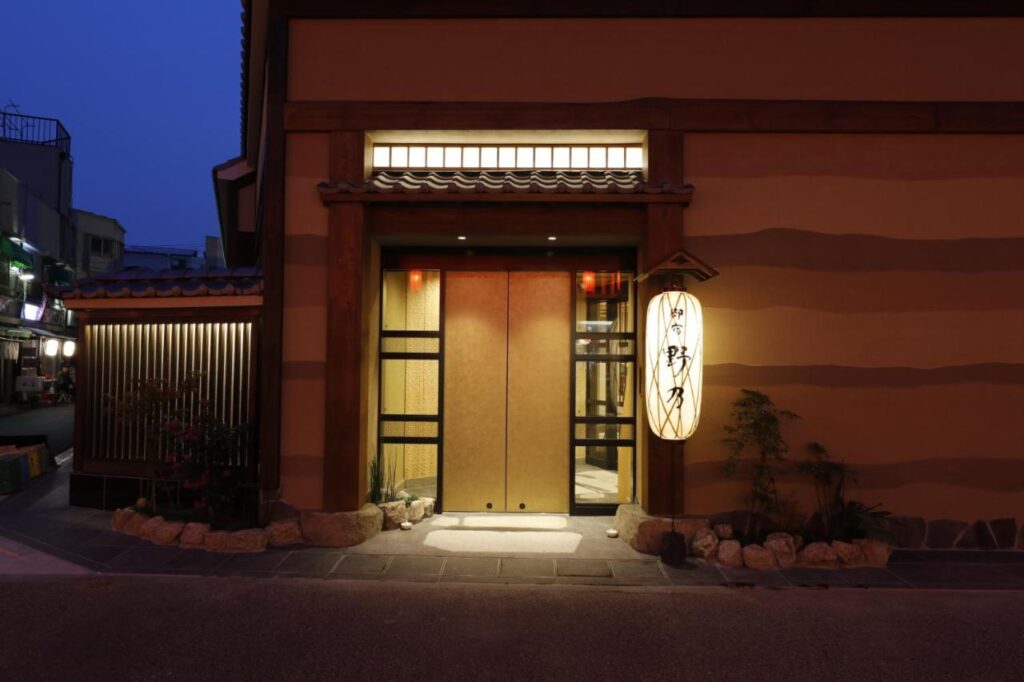 15 Unique Hotels in Tokyo: Coolest hotels 14 A warmly lit entrance to a traditional Japanese building at dusk, featuring sliding wooden doors, a large paper lantern with Japanese writing, and simple landscaping—perfect for those seeking unique hotels in Tokyo.