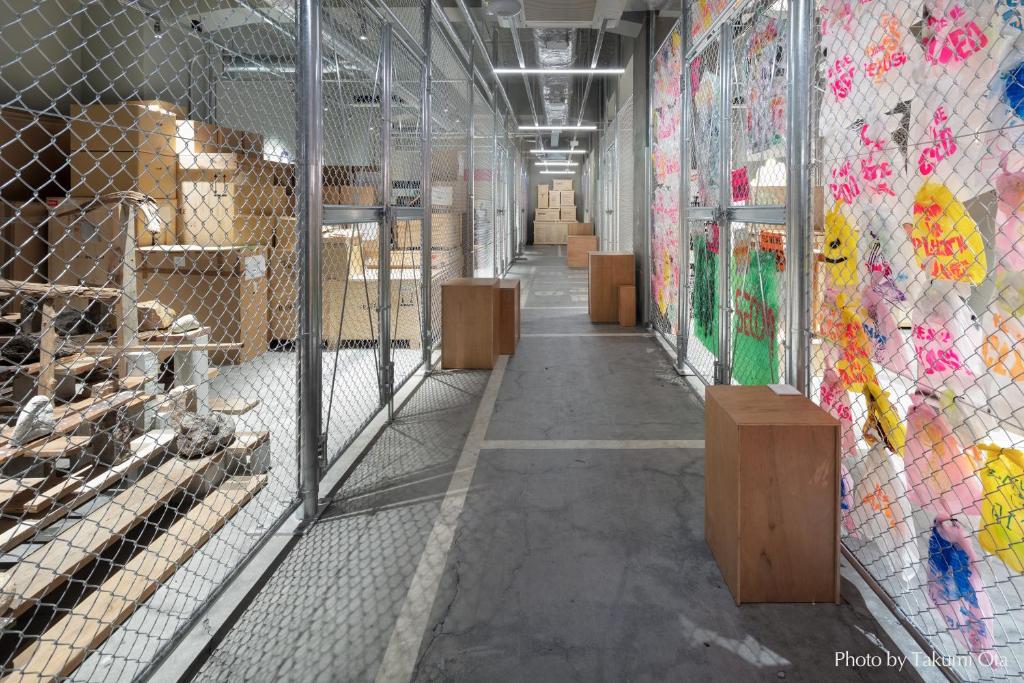 15 Unique Hotels in Tokyo: Coolest hotels 49 A hallway in one of the unique hotels in Tokyo features chain-link fences on both sides, wooden box seating, vibrant translucent panels to the right, and stacked crates with rocks to the left—bright ceiling lights complete the modern vibe.