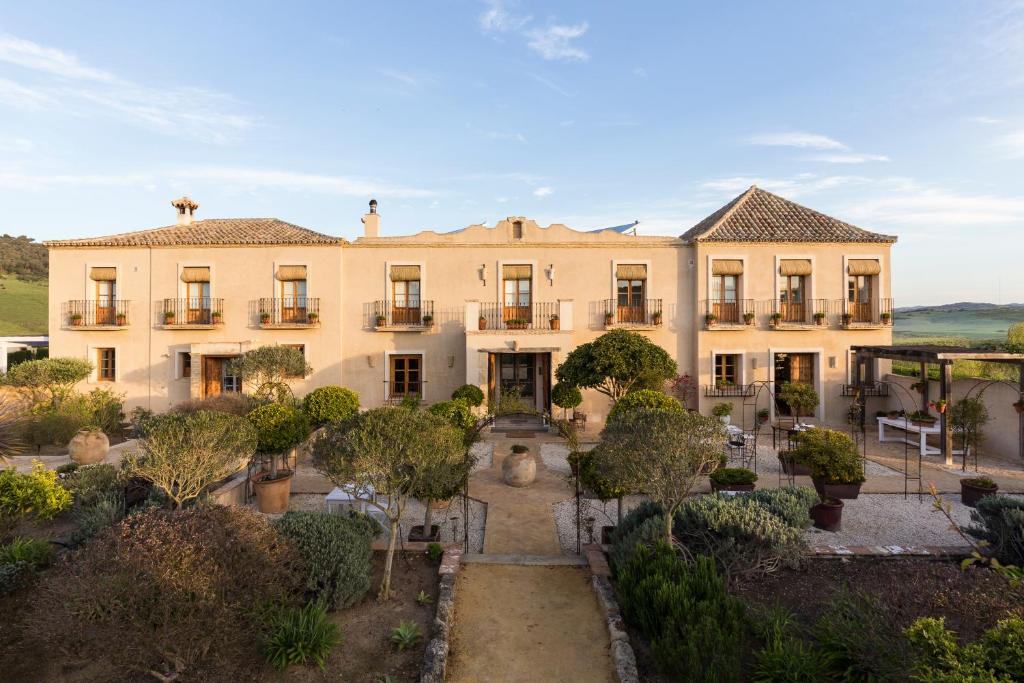 TOP 10 Luxury Hotels in Andalucía 13 A large, two-story beige villa with a tiled roof and balconies resembles the elegance of luxury hotels in Andalucía, surrounded by neatly landscaped gardens and potted plants, set against a backdrop of green hills under a clear blue sky.