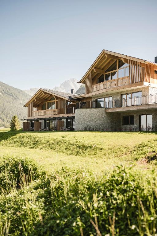 Two modern wooden chalets with large balconies sit on a grassy hillside, surrounded by greenery and distant mountains under a clear, sunny sky—an ideal retreat among the best hotels in the Dolomites.