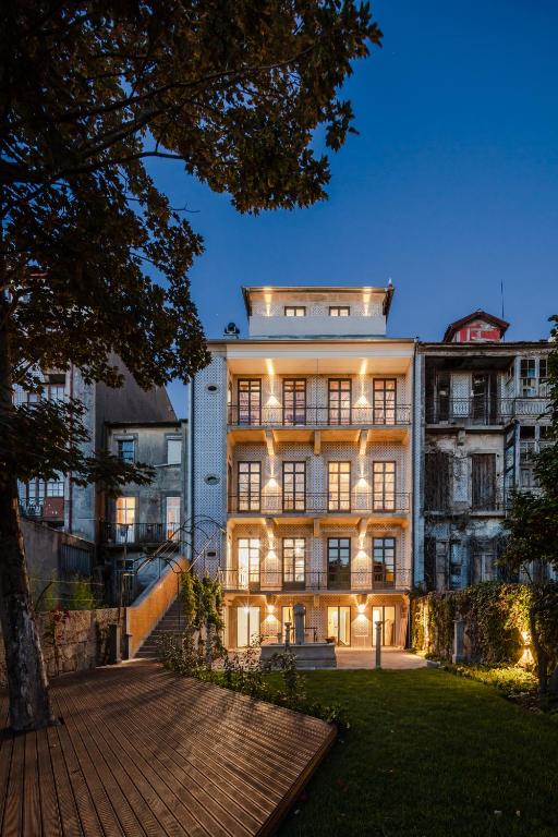 Top 12 Boutique Hotels in Porto, Portugal 28 A four-story building with illuminated balconies at dusk, surrounded by a garden and wooden deck, nestled between older, weathered neighboring structures—reflecting the charm of boutique hotels in Porto.