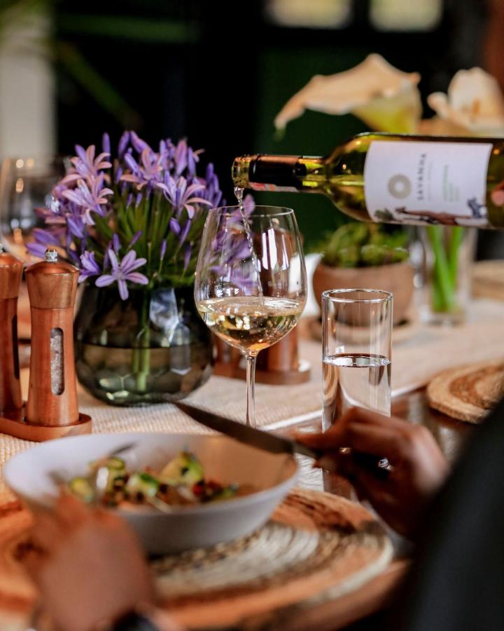 Best Hotels in Rwanda: Unique Hotels and Lodges 3 A person pours white wine into a glass on a table set with salad, water, salt and pepper shakers, and a vase of purple flowers—capturing the warm, inviting atmosphere you’d find at the best hotels in Rwanda.