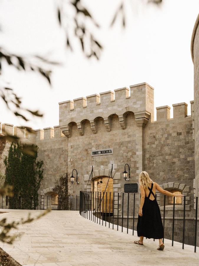 Most Authentic Boutique Hotels in Montenegro, Europe 2 A woman in a black dress walks toward a stone castle entrance with battlements and ivy, under warm sunlight—echoing the charm of boutique hotels in Montenegro. Tree branches frame the enchanting scene.