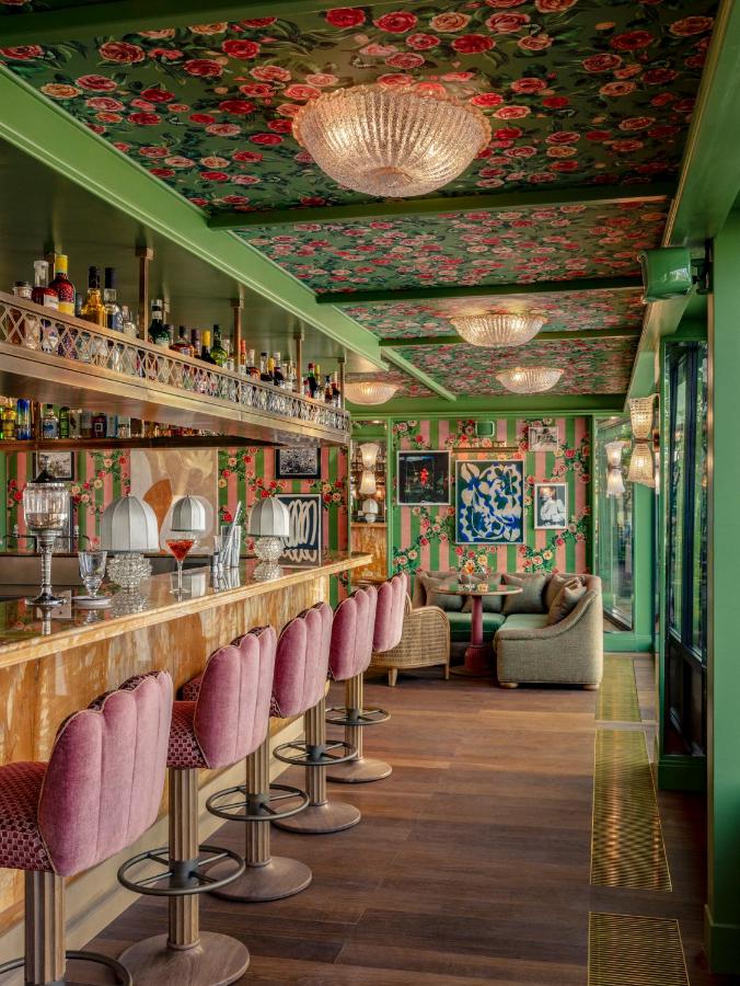 13 Unique Hotels in Paris for a Stylish Stay 6 A vibrant bar in one of the most unique hotels in Paris, featuring pink velvet stools, a marble counter, floral-patterned green walls and ceiling, chandeliers, eclectic artwork, and large windows that create a cozy and colorful interior.