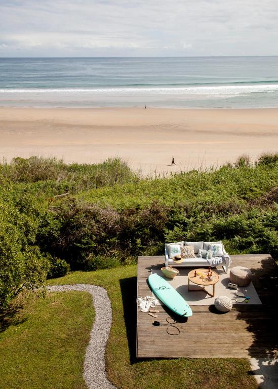 A wooden deck with a sofa, table, cushions, and a turquoise surfboard overlooks a sandy beach and the ocean—an inviting scene reminiscent of boutique hotels in Cantabria, surrounded by lush greenery and a winding path in the foreground.