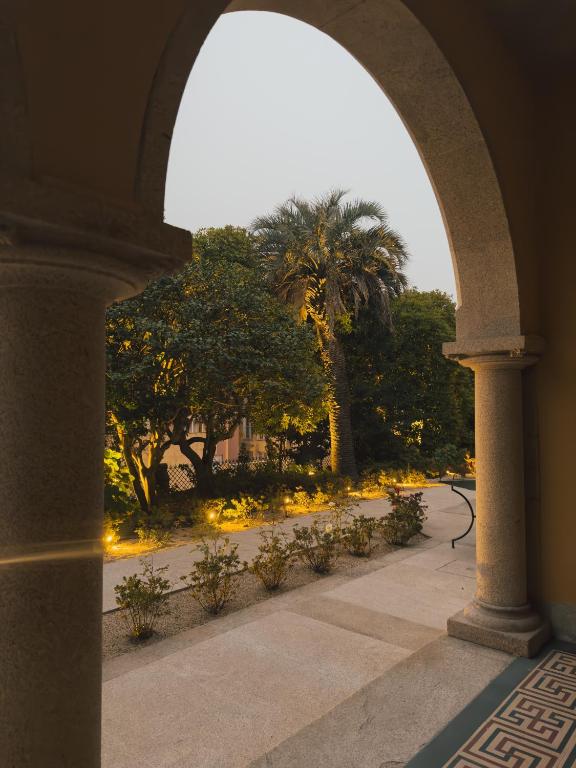 Top 12 Boutique Hotels in Porto, Portugal 51 View through a stone archway onto a garden path lined with bushes and trees, including a tall palm tree, with warm lighting illuminating the plants and a table near the columns—evoking the charm of boutique hotels in Porto.
