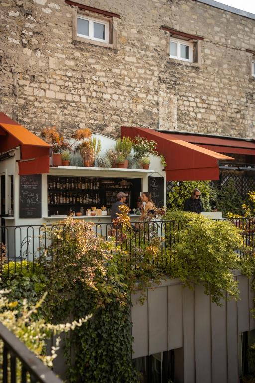 13 Unique Hotels in Paris for a Stylish Stay 54 A small outdoor café with red awnings and shelves of bottles sits on a terrace covered in greenery—an ambiance often found at unique hotels in Paris. Three people are at the counter, with an old stone wall in the background.