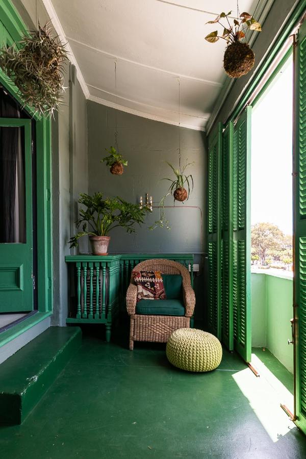 Unique Places to Stay in Cape Town 21 A cozy balcony with green floors, walls, and shutters features a wicker chair with a patterned pillow, a knitted pouf, potted plants, and hanging planters—one of the unique places to stay in Cape Town, all bathed in natural light.