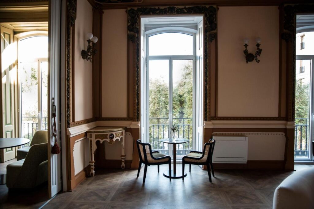 A sunlit room with tall windows, ornate trim, two black chairs, and a small round table—echoing the inviting charm of boutique hotels in Cantabria. A decorative console sits in the corner and trees are visible outside the windows.