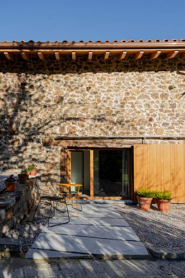 A rustic stone building typical of charming hotels in Cantabria, with a wooden door, large windows, and a cozy patio featuring two chairs, a table, and potted plants. Tree shadows dance across the wall beneath the clear blue sky.