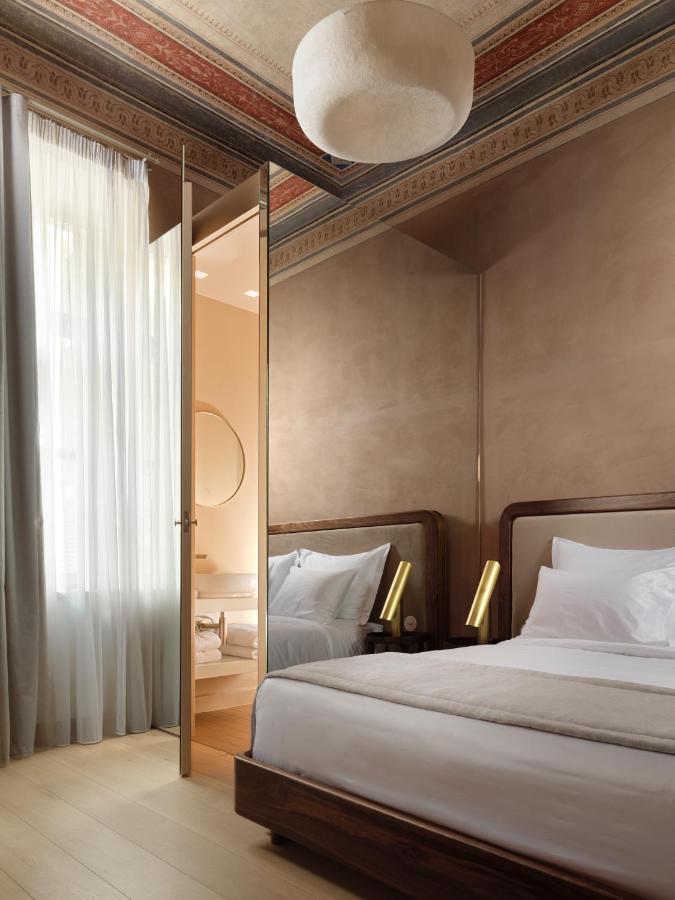 A stylish bedroom reminiscent of the best boutique hotels in Athens, featuring a double bed with white bedding, modern lighting, beige walls, tall curtains, a partially open door to a bathroom with a round mirror, and ornate ceiling details above.