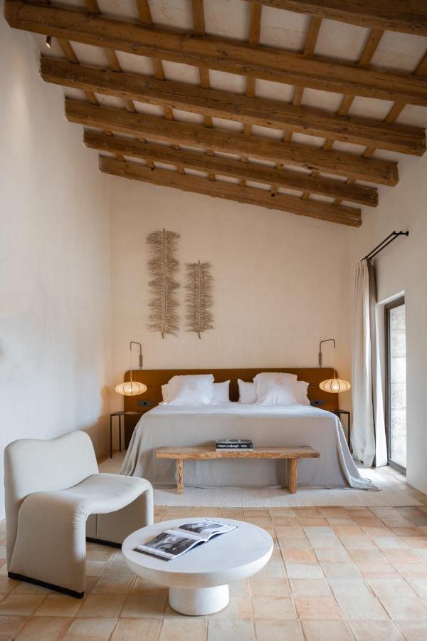 Unique Boutique Hotels in Menorca 3 Minimalist bedroom inspired by boutique hotels in Menorca, featuring a wooden beamed ceiling, a large bed with white linens, hanging lamps, a wooden bench, modern white chair, and a round coffee table on terracotta tile flooring.