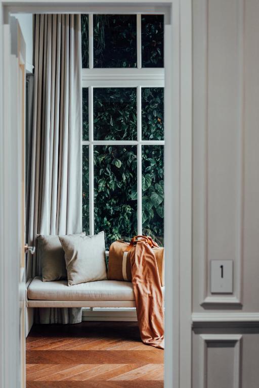 A cozy window seat with beige cushions and a burnt orange throw blanket, set beside tall curtains and large windows overlooking lush greenery—reminiscent of the inviting nooks found in boutique hotels in Athens—seen through an open doorway.