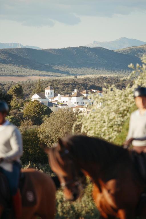 TOP 10 Luxury Hotels in Andalucía 23 Two people on horseback ride through a sunlit landscape with greenery in the foreground and a luxury hotel in Andalucía nestled among trees, hills, and mountains in the background.