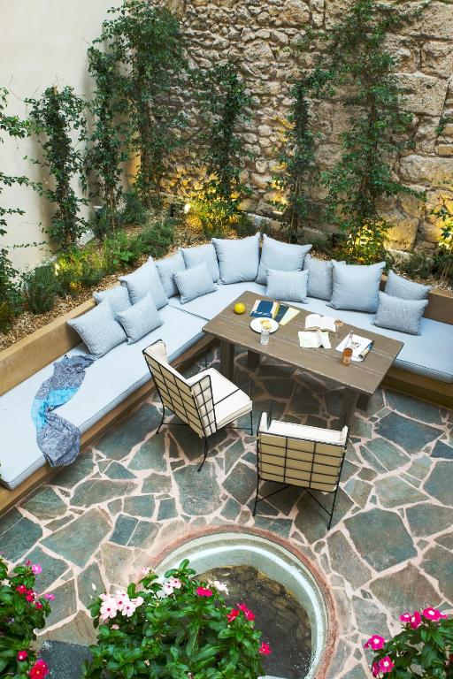 Cozy outdoor patio with a stone floor, a circular water feature, L-shaped cushioned bench, two chairs, a table with books and drinks—evoking the charm of boutique hotels in Athens—surrounded by greenery and flowering plants against a stone wall.