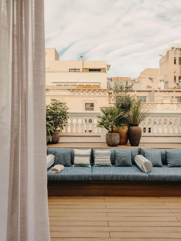 13 Unique Hotels in Barcelona, Spain 28 A cozy outdoor terrace at one of the unique hotels in Barcelona features a long blue cushioned sofa, patterned pillows, potted plants, wooden flooring, and city views beneath a partly cloudy sky. Sheer curtains hang on the left side.