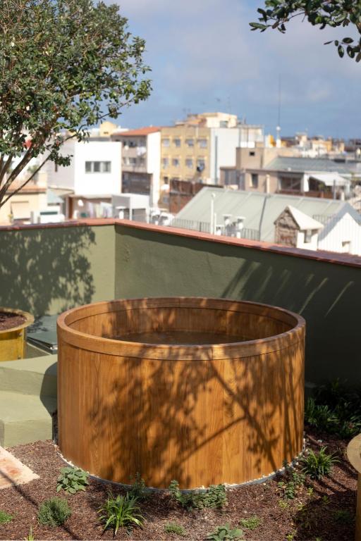 13 Unique Hotels in Barcelona, Spain 52 A wooden round hot tub sits on a rooftop garden surrounded by small plants and trees, with city buildings visible in the background under a partly cloudy sky—perfect for guests seeking unique hotels in Barcelona.