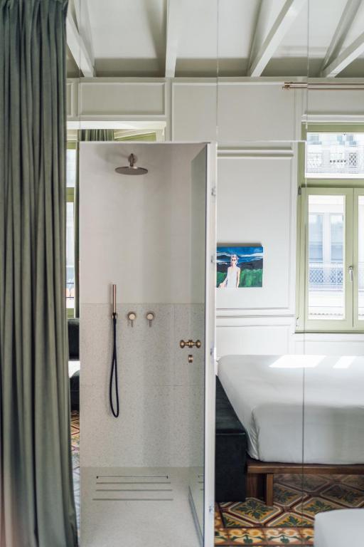 A modern bedroom inspired by boutique hotels in Athens features a glass shower stall, green curtains, a bed with white linens, large windows with green frames, and a wall painting. Natural light highlights the patterned floor tiles.