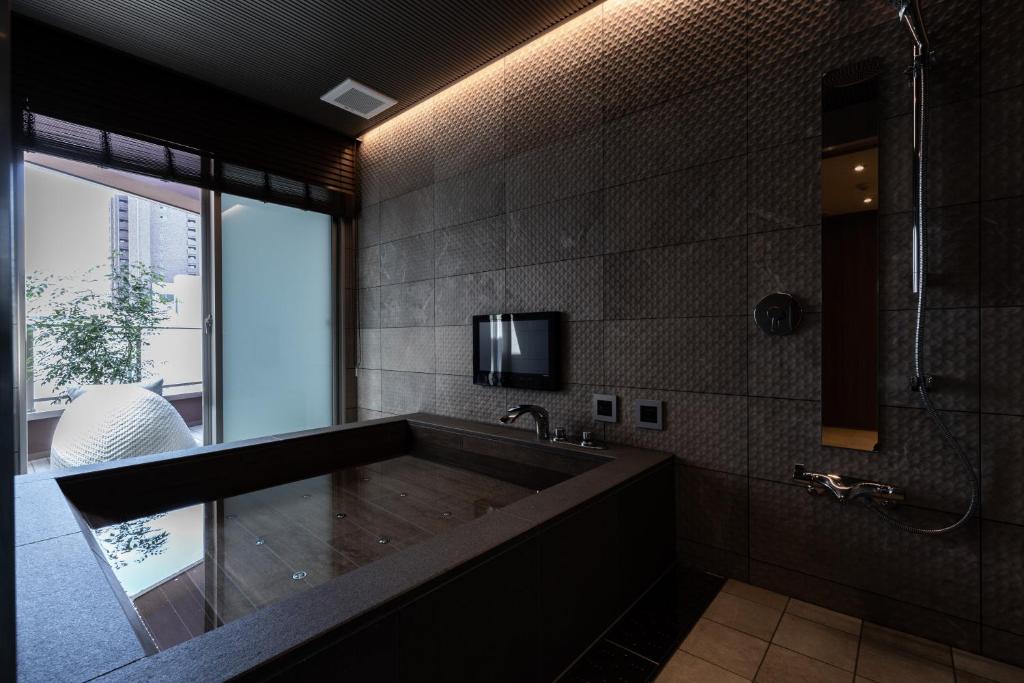 7 Best Hotels in Hiroshima, Japan 29 Modern bathroom at one of the best hotels in Hiroshima, featuring a dark stone soaking tub, wall-mounted TV, large frosted window, rainfall shower, and round white décor by a potted plant. Warm ambient lighting highlights textured walls.